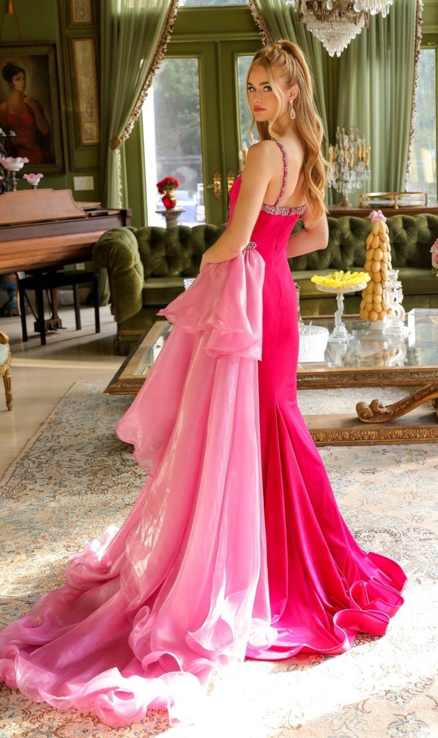 A woman in the Long Prom Dress 39556 by Ava Presley—a bright pink fitted gown with a plunging v-neckline and long train—stands in a luxurious living room featuring green walls, a chandelier, piano, and ornate furniture.